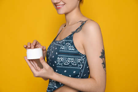 Woman with tattoo holding jar of cream against yellow background, closeupの写真素材