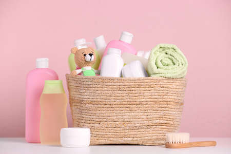Baby cosmetic products, bath accessories and toy in wicker basket on white table against pink...