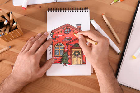 Man drawing in sketchbook with pencil at wooden table, top view