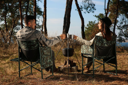 Couple resting in camping chairs and enjoying hot drink near bonfire outdoorsの写真素材