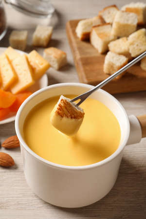 Dipping piece of bread into tasty cheese fondue on light wooden tableの写真素材