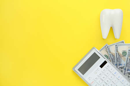 Ceramic model of tooth, dollar banknotes and calculator on yellow background, flat lay with space for text. Expensive treatmentの写真素材