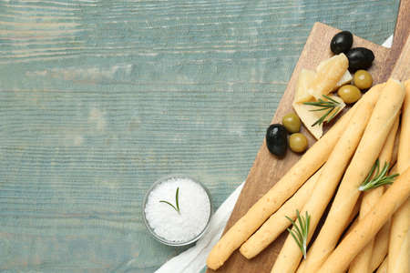 Tasty grissini with rosemary, cheese, olives and salt on light blue wooden table, flat lay. Space...