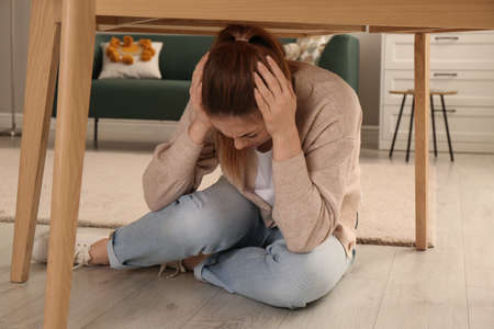 Scared woman hiding under table in living room during earthquakeの写真素材