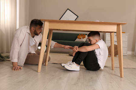 Father comforting his scared son under table in living room during earthquakeの写真素材