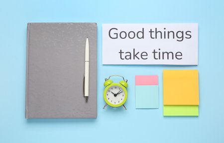 Different stationery, alarm clock and card with motivational quote Good Things Take Time on light blue background, flat layの写真素材