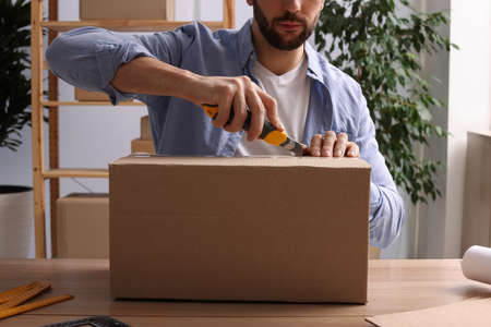 Man using utility knife to open parcel at wooden table indoors, closeupの写真素材