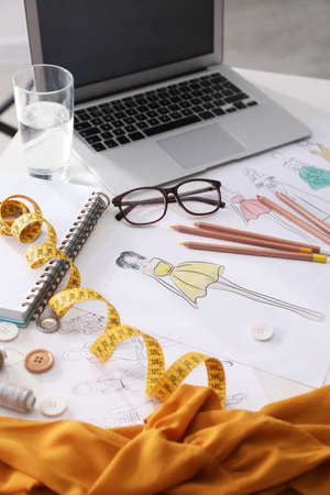 Sketches of clothes and different stuff on white table indoors. Fashion designer's workplace