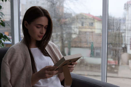 Melancholic young woman with photo frame sitting in armchair indoors, space for text. Loneliness conceptの写真素材