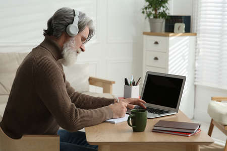 Middle aged man with laptop, notebook and headphones learning at table indoors