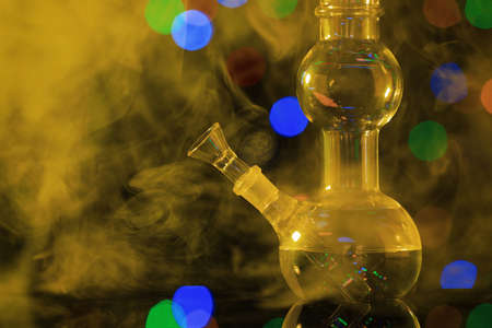 Closeup view of glass bong with smoke against blurred lights, toned in yellow. smoking device