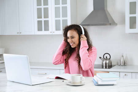 African American woman with modern laptop and headphones studying in kitchen. distance learning