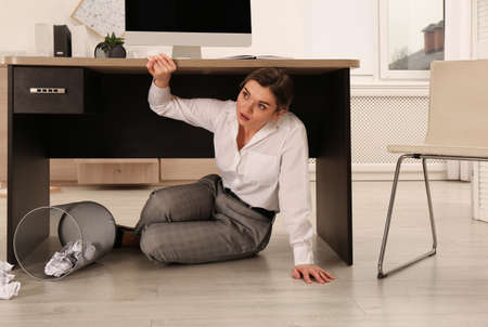 Scared young woman hiding under office desk during earthquakeの写真素材