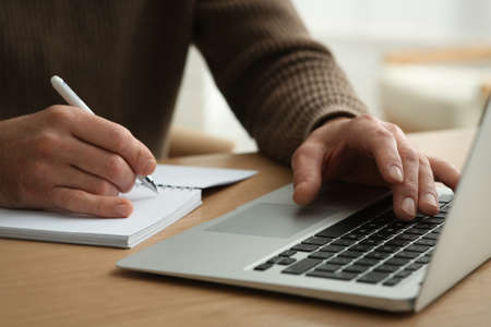 Man with laptop and notebook learning at wooden table indoors, closeup