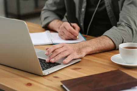 Man with laptop and notebook learning at wooden table indoors, closeup