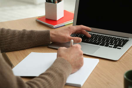 Man with laptop and notebook learning at wooden table indoors, closeup