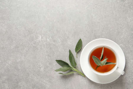 Cup of aromatic sage tea and fresh leaves on gray table, flat lay. Space for text