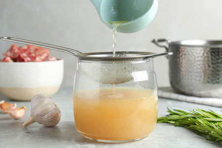 Sieving delicious bone broth into glass jar on light gray table