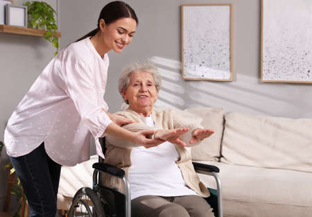 Senior woman in wheelchair doing physical exercise and young caregiver helping her indoors. home health care serviceの写真素材