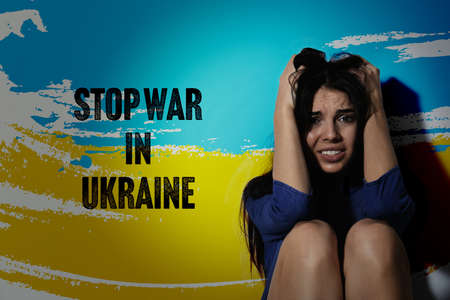 Stop War in Ukraine. Afraid young woman sitting near wall painted in colors of Ukrainian national flagの写真素材