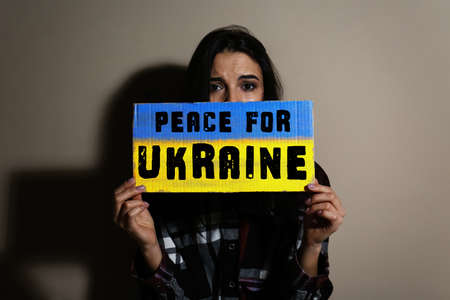 Sad woman holding sign with phrase Peace for Ukraine near beige wallの写真素材