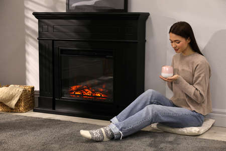 Beautiful young woman with cup of hot drink sitting on floor near fireplace at home. Space for textの写真素材