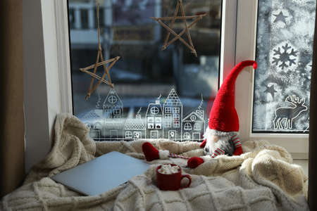 Gnome, laptop and hot drink on knitted plaid near window with beautiful drawing at home. christmas...