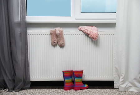 Heating radiator with knitted hat, socks and rubber boots near window indoorsの写真素材