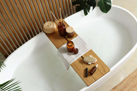 Wooden bath tray with open book, candle and body care products on tub indoors. Relaxing atmosphereの写真素材