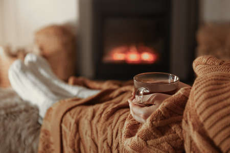 Woman with cup of tea resting near fireplace at home, closeupの写真素材