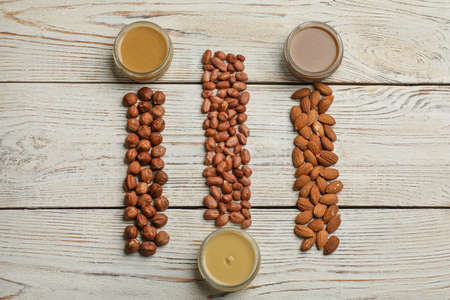 Jars with butters made of different nuts and ingredients on white wooden table, flat lay