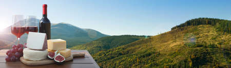 Tasty wine, grapes and cheeses on wooden table against beautiful mountain landscape, space for text. banner designの写真素材