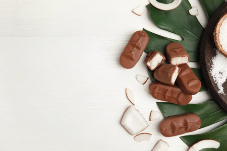 Delicious milk chocolate candy bars with coconut filling on white wooden table, flat lay. Space for textの写真素材