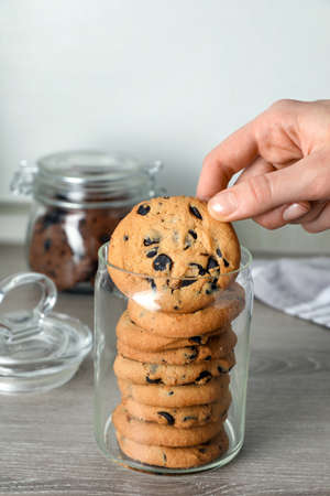 Woman taking delicious chocolate chip cookie from glass jar at wooden table, closeupの写真素材