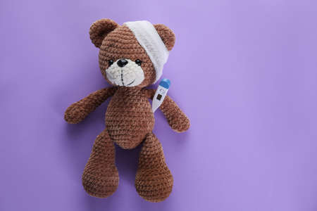 Toy bear with bandage and thermometer on purple background, top view with space for text. children's...