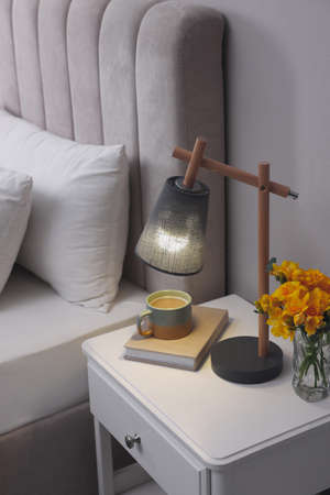 Stylish lamp, book, hot drink and flowers on bedside table indoors. bedroom interior elementの写真素材