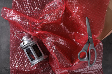 Beautiful decorative lantern, red bubble wrap, scissors and cardboard box on dark gray table, flat...