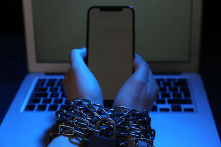 Woman with chained hands holding smartphone near laptop on dark background, closeup. internet...