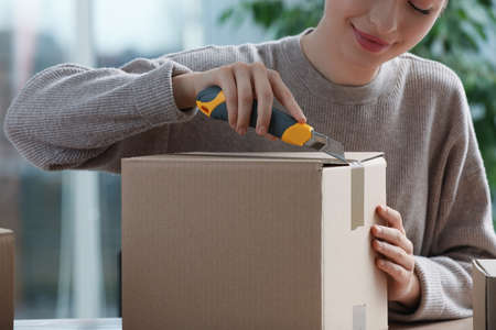 Young woman using utility knife to open parcel indoors, closeupの写真素材