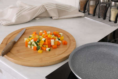 Wooden board with cut vegetables and knife near frying pan in kitchenの写真素材