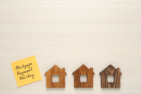Paper note with words Mortgage Payment Holiday and house models on white wooden table, flat lay....