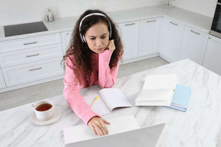 African American woman with modern laptop and headphones studying in kitchen. distance learning