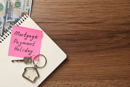 Note with words Mortgage Payment Holiday, key, notebook and money on wooden table, flat lay. Space...