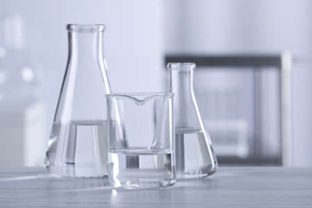 Different laboratory glassware with transparent liquid on wooden table against blurred background....