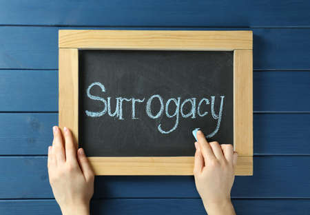 Woman writing word Surrogacy on small blackboard at blue wooden table, top viewの写真素材