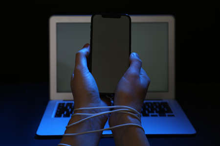 Woman with tied hands holding smartphone near laptop on dark background, closeup. internet addiction