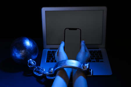 Woman shackled with ball and chain holding smartphone near laptop on dark background, closeup. internet addictionの写真素材
