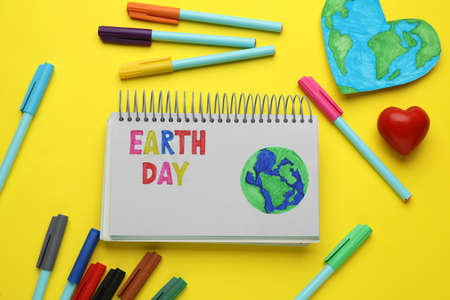 Notebook with phrase Earth Day, heart shaped model of planet and felt pens on yellow background, flat layの写真素材