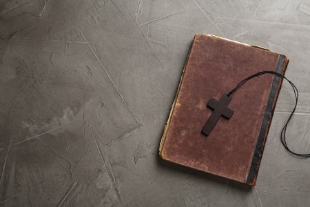 Wooden Christian cross and old Bible on gray table, top view. Space for text
