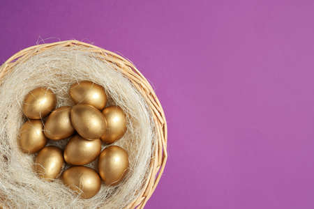 Shiny golden eggs in nest on violet background, top view. Space for textの写真素材